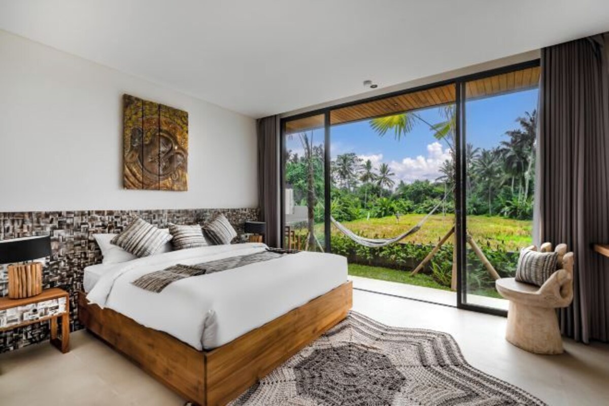 A bedroom with a large bed, a view of a lush garden, and a hammock outside the window.