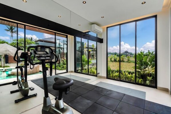 Une salle de sport moderne, dotée d’équipements d’exercice et de grandes fenêtres donnant sur une piscine et une végétation luxuriante.