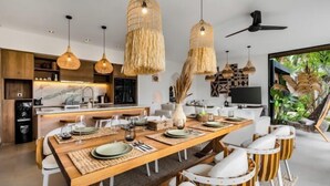 Una cocina moderna con salpicadero de mármol, armarios de madera y una zona de comedor con una mesa de madera alargada y sillas blancas.