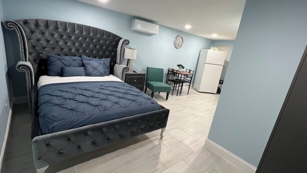 A bedroom with a large bed, a desk, a chair, a small table, and a refrigerator.