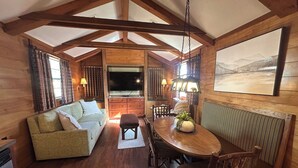 Interior acogedor de una cabaña con techo de madera, un sofá, una mesa de comedor con sillas y un amplio ventanal con cortinas.