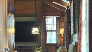 Un interior rústico con vigas de madera, una zona de comedor con sillas y una mesa, y una ventana con cortinas.
