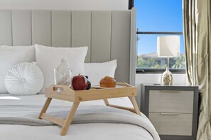 Cama bem arrumada com uma bandeja de madeira com um muffin e uma maçã, mesa de cabeceira com abajur e vista para o verde lá fora.