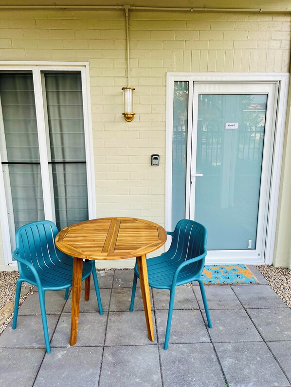 Une terrasse avec une table ronde en bois et deux chaises bleues, une porte-fenêtre coulissante et un mur en briques.