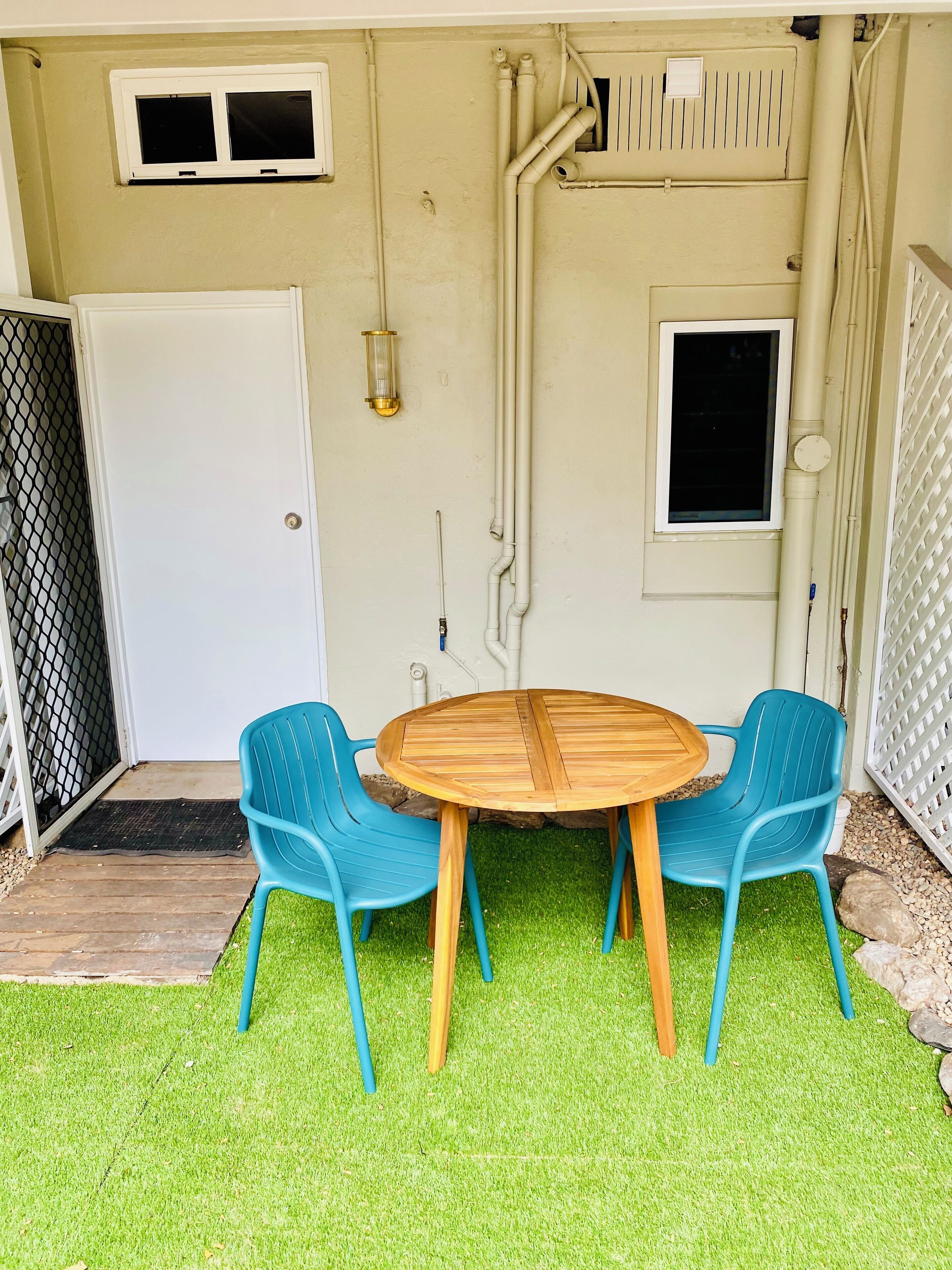 Un pequeño patio con una mesa redonda de madera y dos sillas de plástico azules, rodeado de césped artificial.