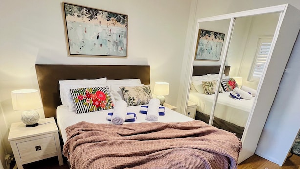 A neatly made bed with a brown blanket, a bedside table with a lamp, and a large mirror. - City Unit 5 Walk to Esplanade (Cairns North)
