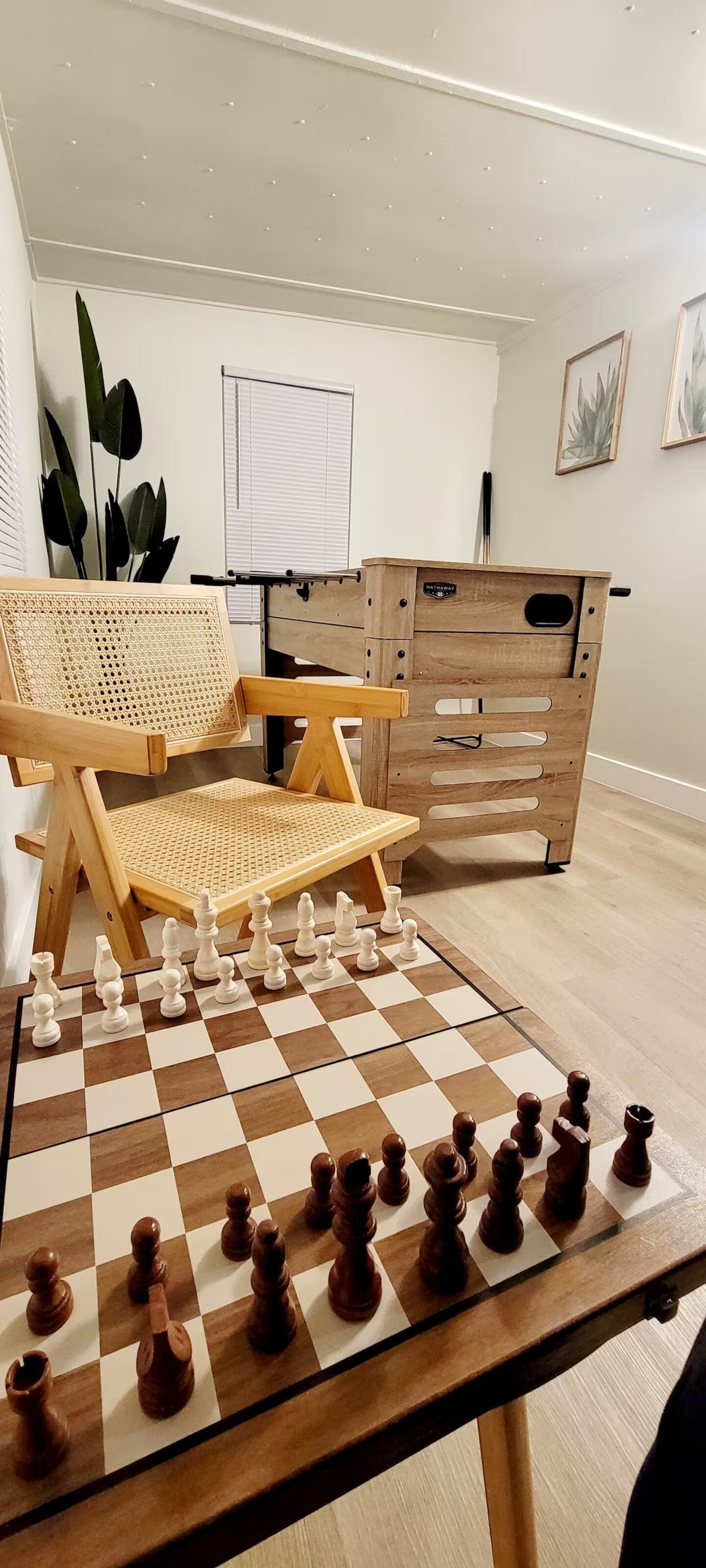 A chessboard set up on a table in a room with a wooden cabinet, a rattan chair, and a window with blinds.