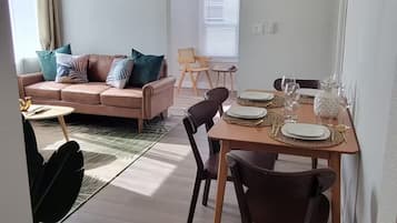A modern living room with a sofa, coffee table, and dining table set for four. A ceiling fan is visible, and the room has a large window.