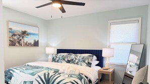 Ein Schlafzimmer mit einem Bett, zwei Nachttischen, einem Deckenventilator, einem Fenster und einem an der Wand befestigten Spiegel.