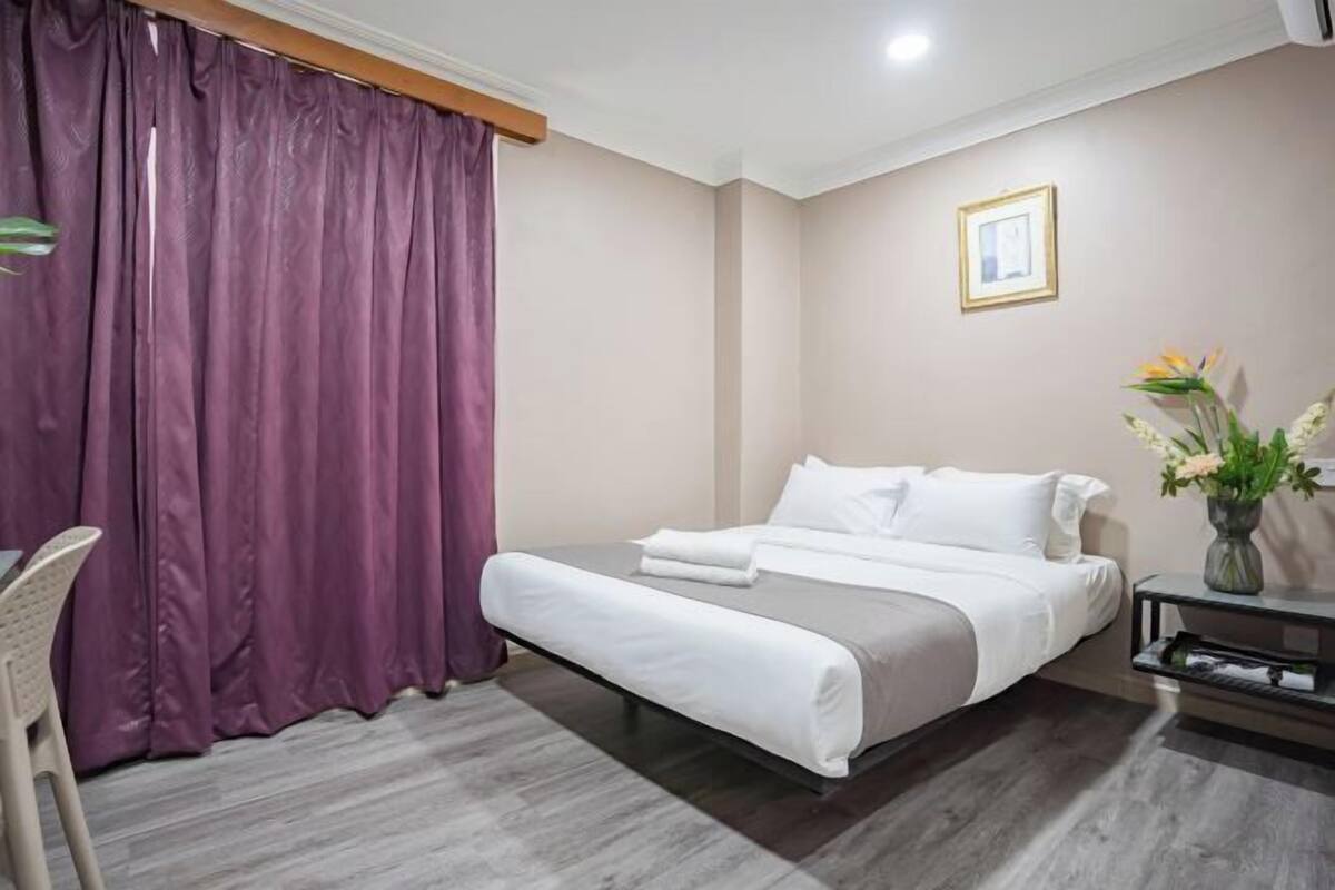 A hotel room with a bed, a desk, a chair, a vase with flowers, and a framed picture on the wall.