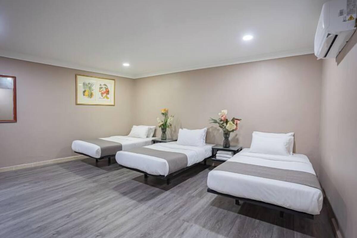 A hotel room with three single beds, a small table with a vase of flowers, and a framed picture on the wall.