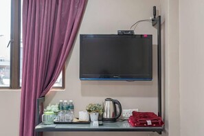 A hotel room with a flat-screen TV mounted on the wall, a small table with a kettle, a red towel, and toiletries.