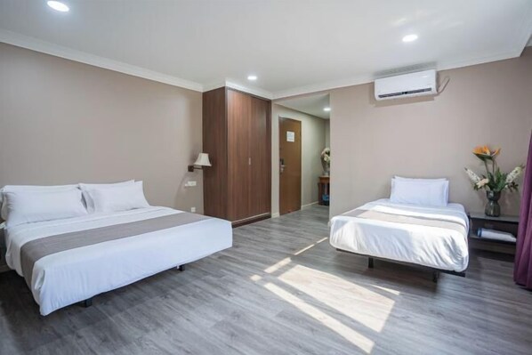 A hotel room with two beds, a wooden wardrobe, a mirror, an air conditioning unit, and a vase with flowers.