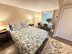 Un dormitorio con cama, mesita de noche, lámpara, escritorio, silla y ventana con vista a árboles.