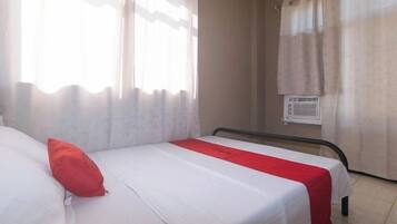 A single bed with a white and red bedspread, a red pillow, and a window with sheer curtains.