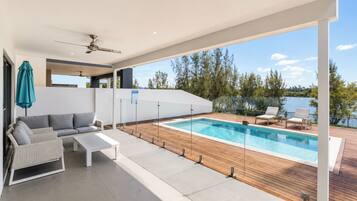 A modern outdoor pool area with a wooden deck, a seating area with a sofa and chairs, and a view of a lake.