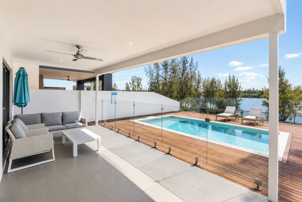 Een modern buitenzwembad met een houten terras, een zithoek met een bank en stoelen, en uitzicht op een meer.