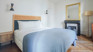 A bedroom with a bed, a wooden nightstand, a wall-mounted lamp, a fireplace, and a large mirror. - Blueground | Saint Vincent de Paul, view, nr Canal (Paris)