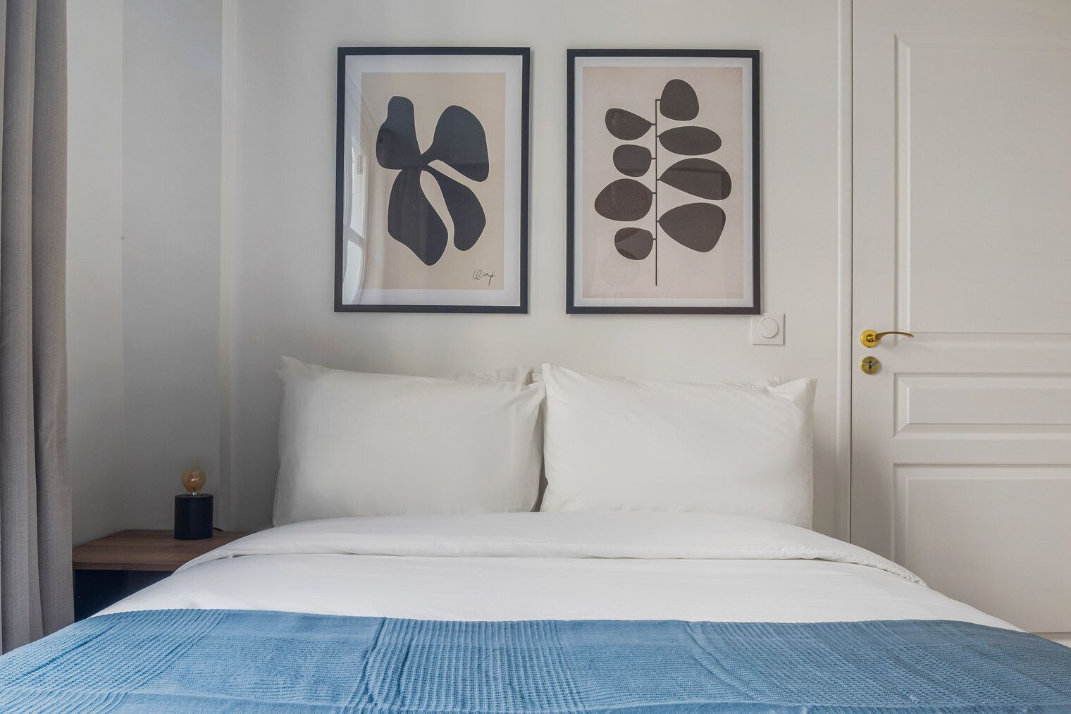A bedroom with a bed, two framed abstract art pieces on the wall, a nightstand, and a door.