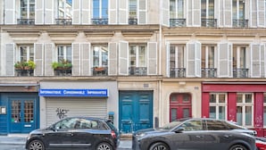 A row of classic European buildings with shutters, a blue storefront, and two parked cars.