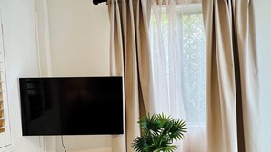 Een moderne woonkamer met een flatscreen televisie die aan een wit kastje is bevestigd, een potplant op het kastje en beige gordijnen voor een raam.