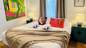 Een slaapkamer met een bed, twee nachtkastjes, een wandairconditioner, een raam met gordijnen en een schilderij van een tropisch huis.