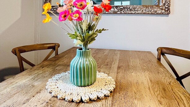 Een houten eettafel met een groen gestreepte vaas met kleurrijke bloemen, een witte tafelonderzetter en twee houten stoelen.