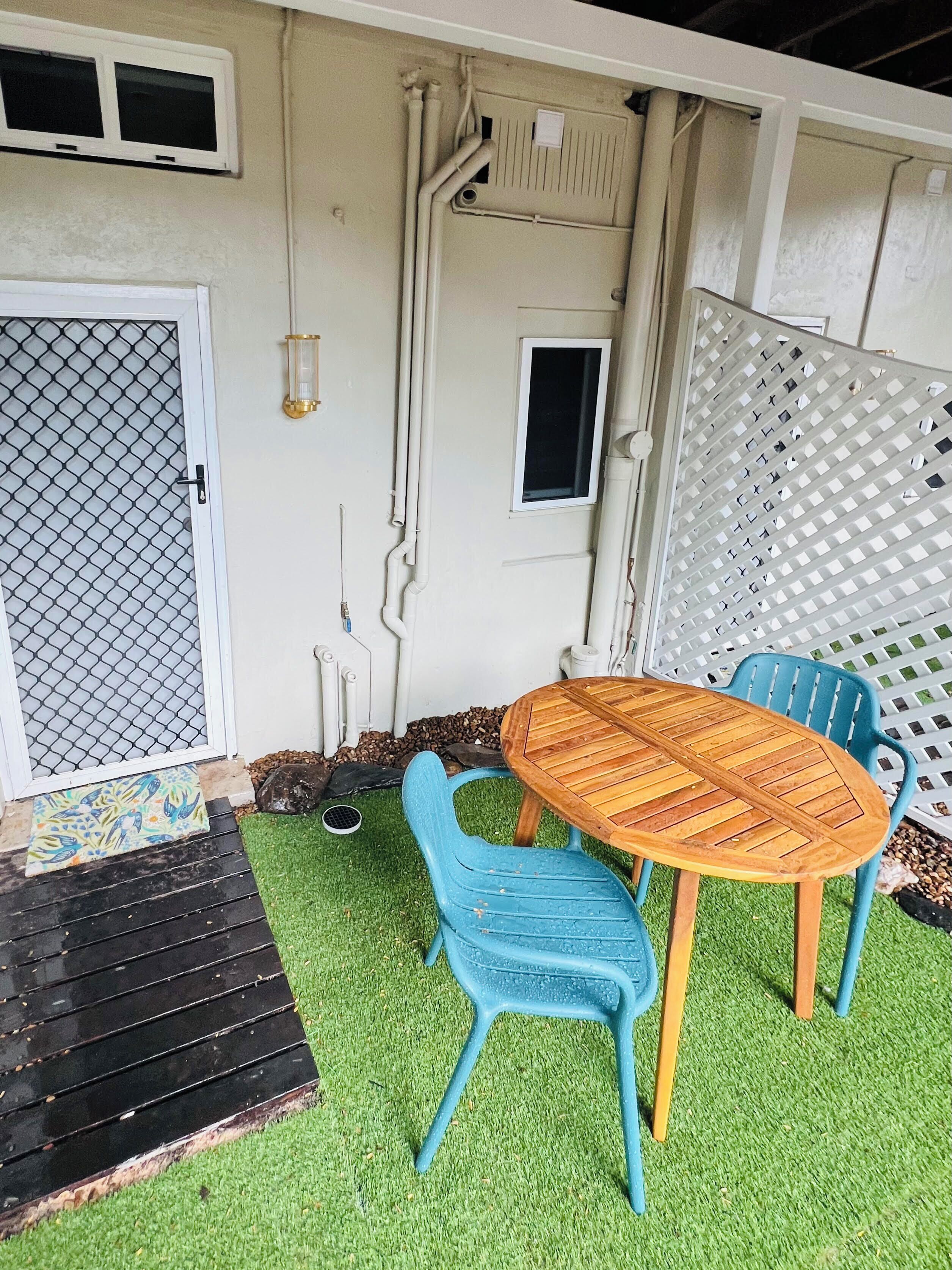 A patio with a wooden table and blue chairs, surrounded by artificial grass.