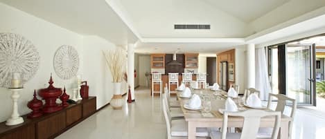 A spacious dining area with a long table set for a meal, a kitchen in the background, and a large window providing natural light.