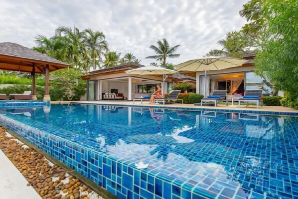 A swimming pool with tiled blue edges, lounge chairs, umbrellas, and a covered seating area. - Baan Ban Buri (Koh Samui)