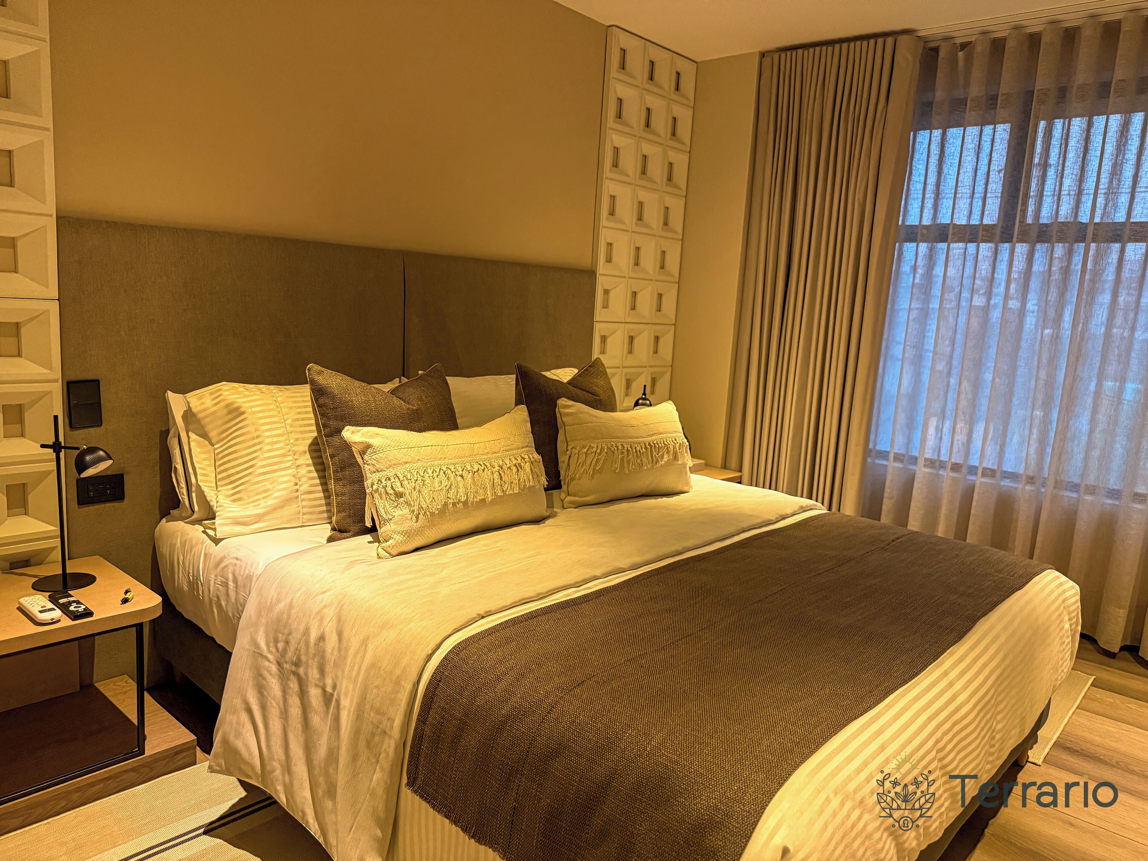 A hotel room with a large bed, a nightstand with a lamp, and a view of the outdoors through the window.