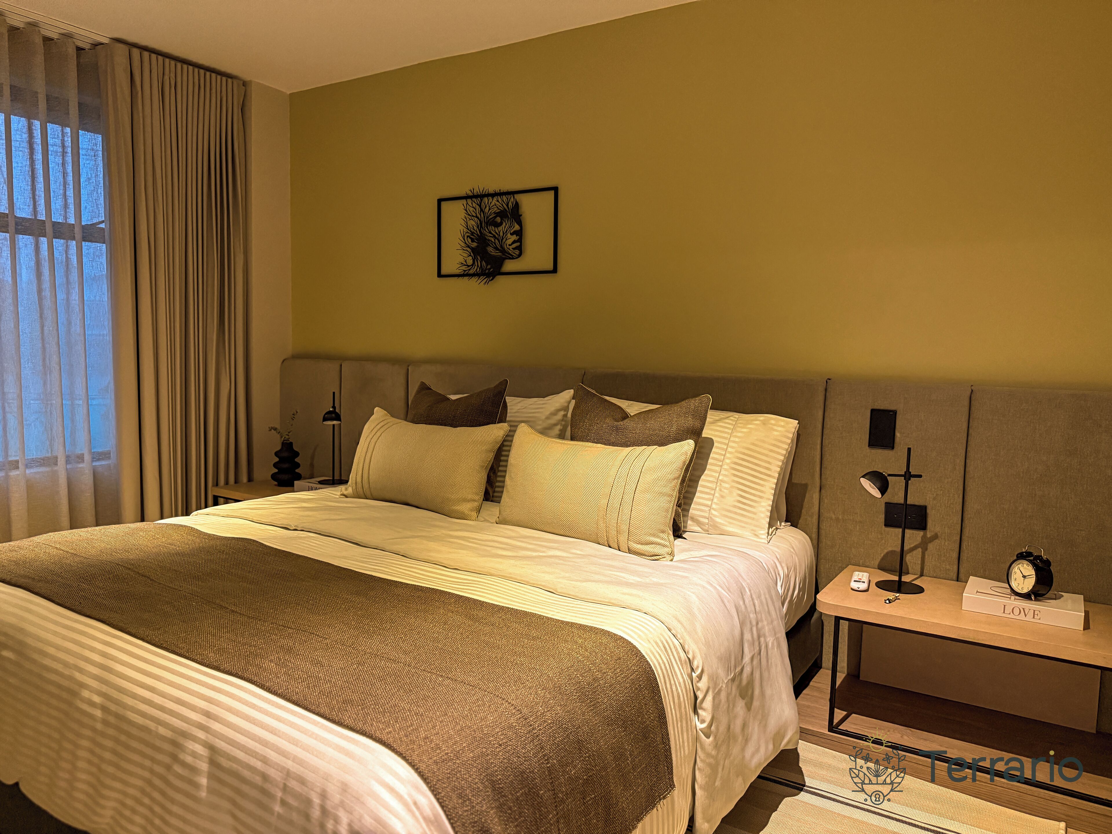 Een net opgemaakt bed met een gestreept dekbed, een nachtkastje met een lamp en een klok, en een ingelijst kunstwerk aan de muur.