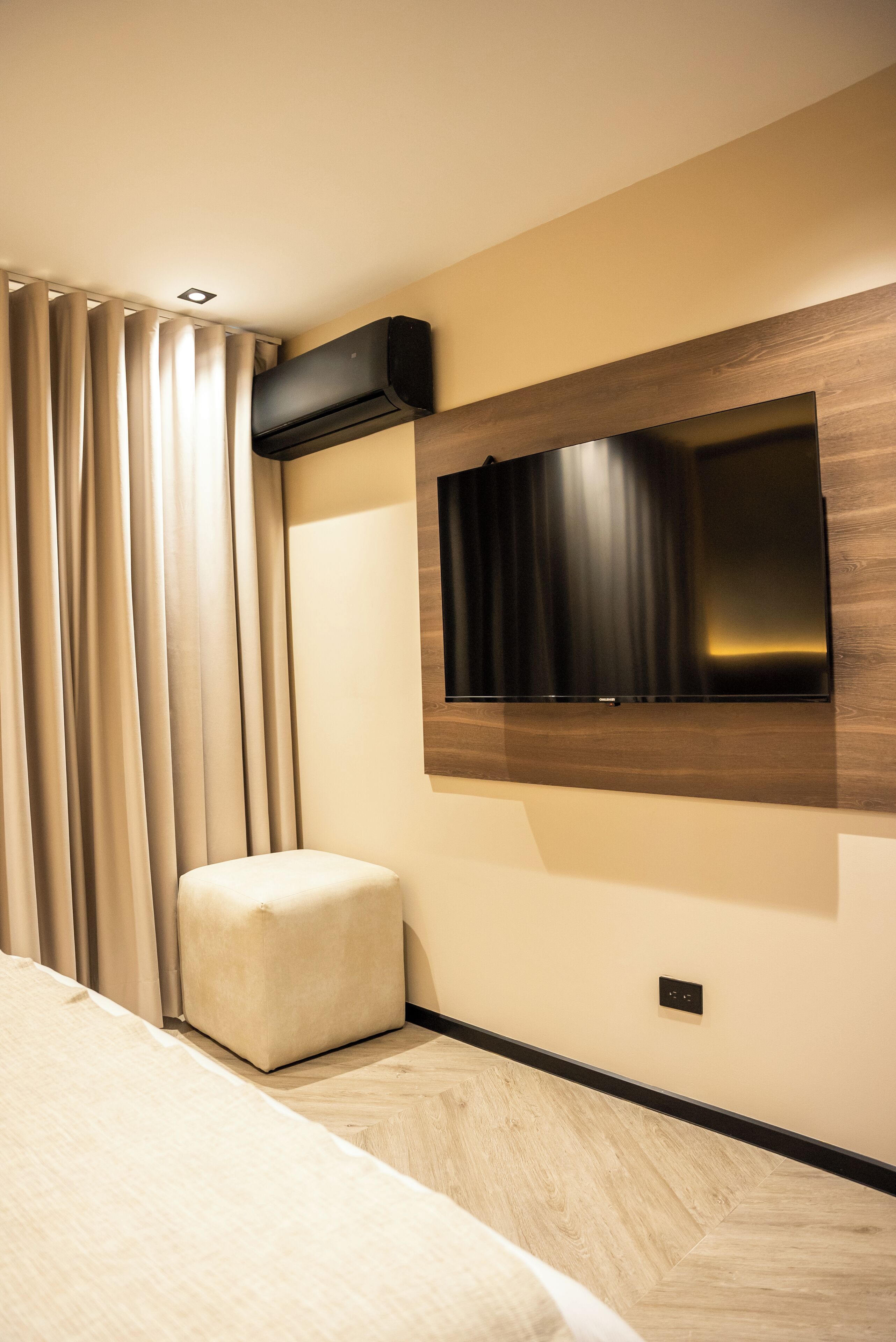A modern hotel room with a flat-screen TV mounted on the wall, a small ottoman, and a ceiling-mounted air conditioning unit.
