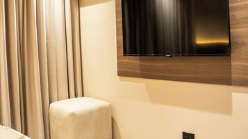 A modern hotel room with a flat-screen TV mounted on the wall, a small ottoman, and a ceiling-mounted air conditioning unit.