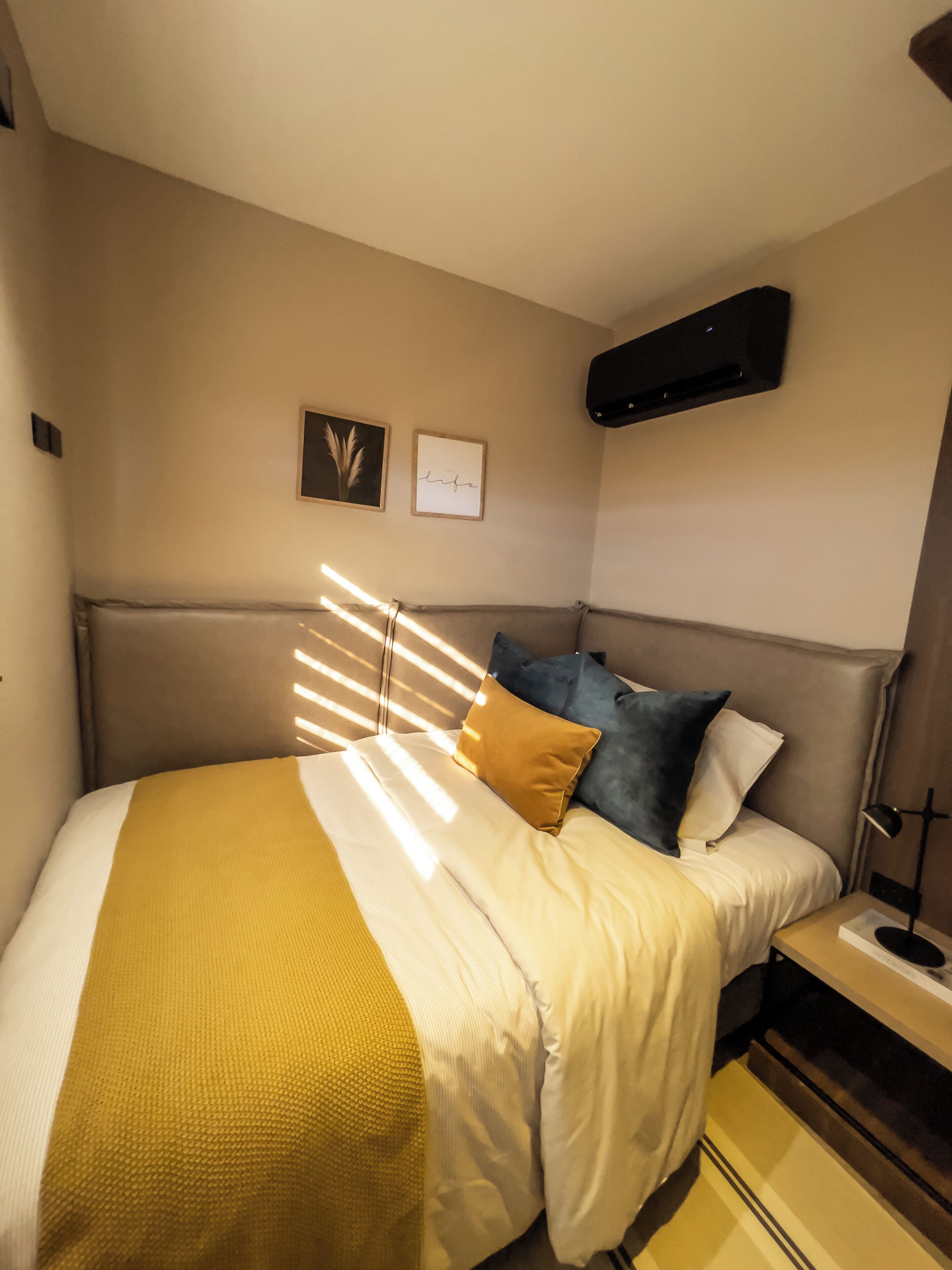 Un lit bien fait, recouvert d’une couverture jaune, avec un oreiller marron et un oreiller bleu. Deux tableaux encadrés sont accrochés au mur, l’un représentant une plante et l’autre portant le mot « vie ».
