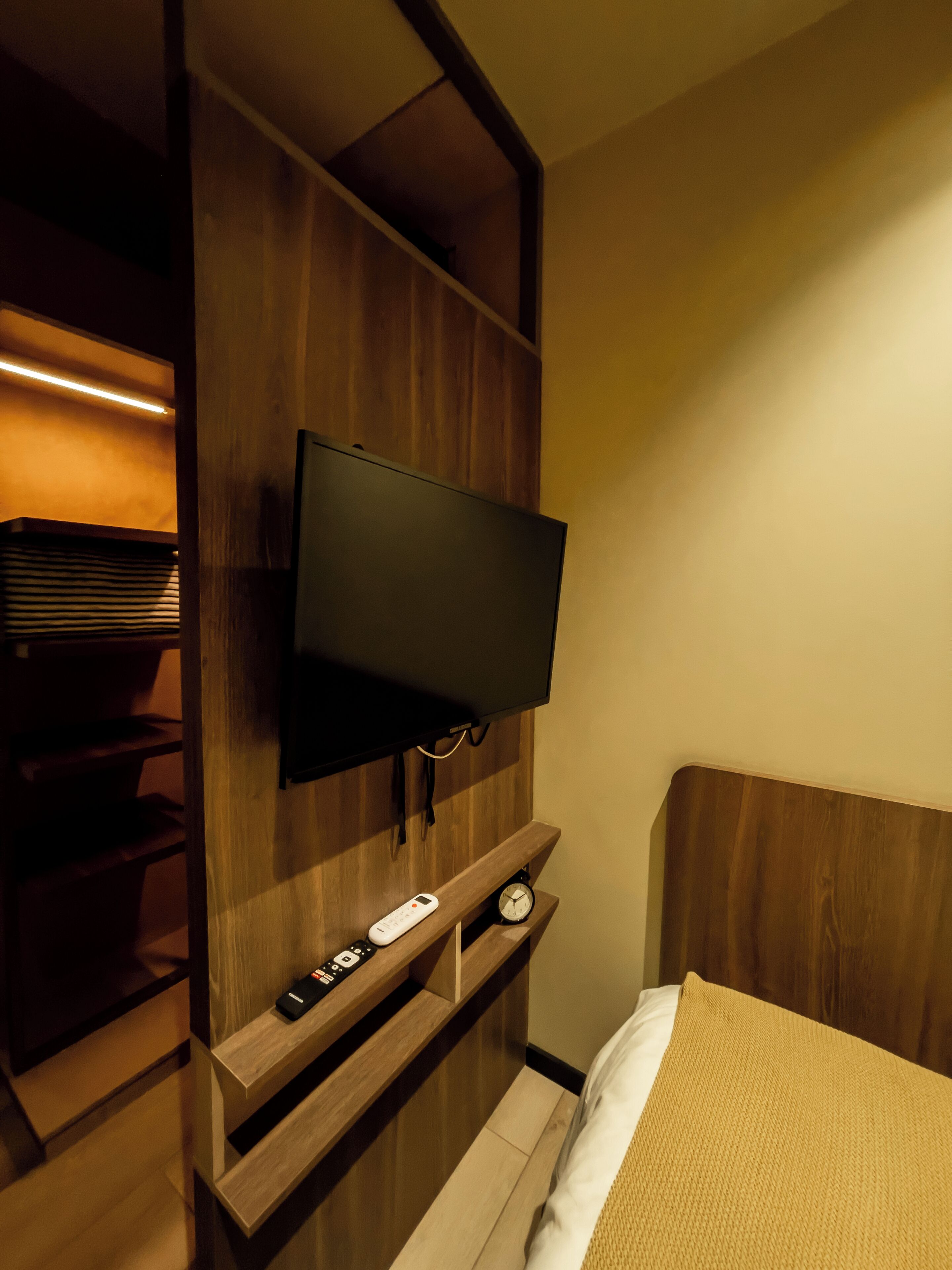 Habitación de hotel con televisor de pantalla plana montado en una pared de madera, una mesita de noche con un reloj y una cama con una manta color beige.