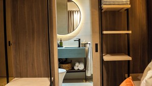 A modern hotel room with a wooden wardrobe, a bed with white bedding and an orange pillow, a bathroom with a mirror, sink, and towel rack. - Terrario las acacias  (Armenia)