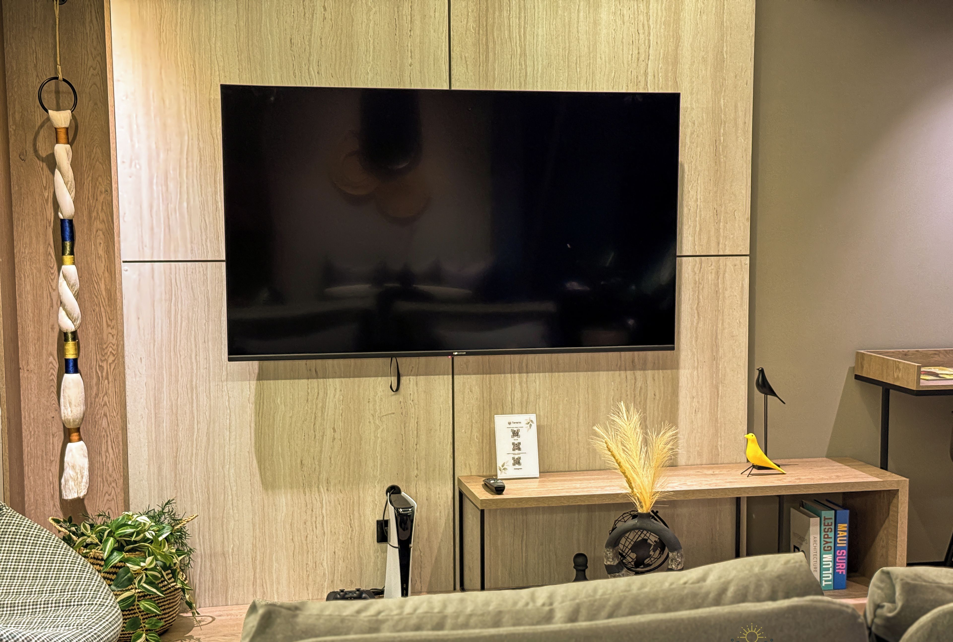 Een moderne woonkamer met een flatscreen televisie aan de muur, een houten console met decoratieve voorwerpen, een groene bank en een fauteuil met een patroon.