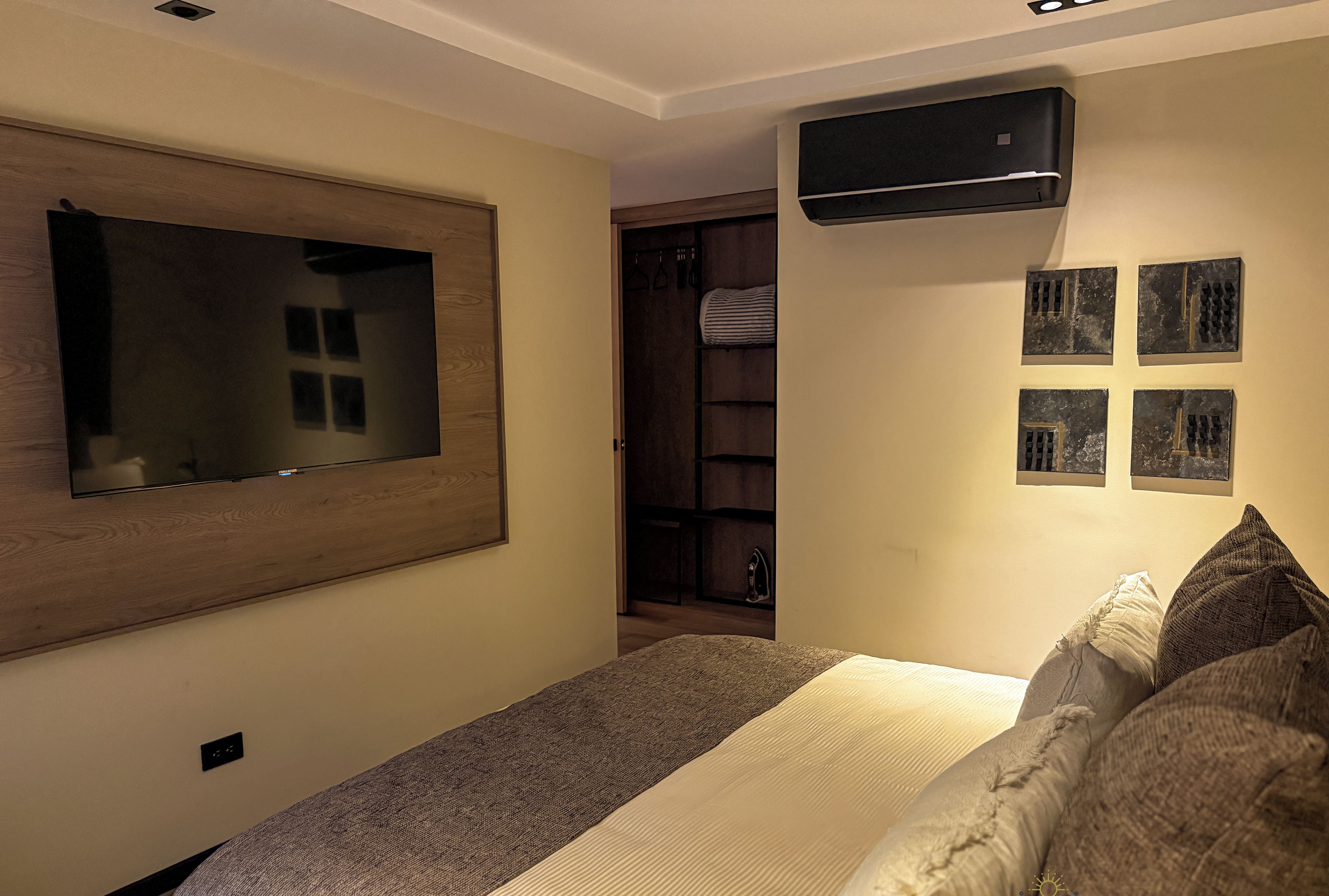 A bedroom with a large bed, a flat-screen TV mounted on the wall, a wardrobe, and a wall-mounted air conditioner.