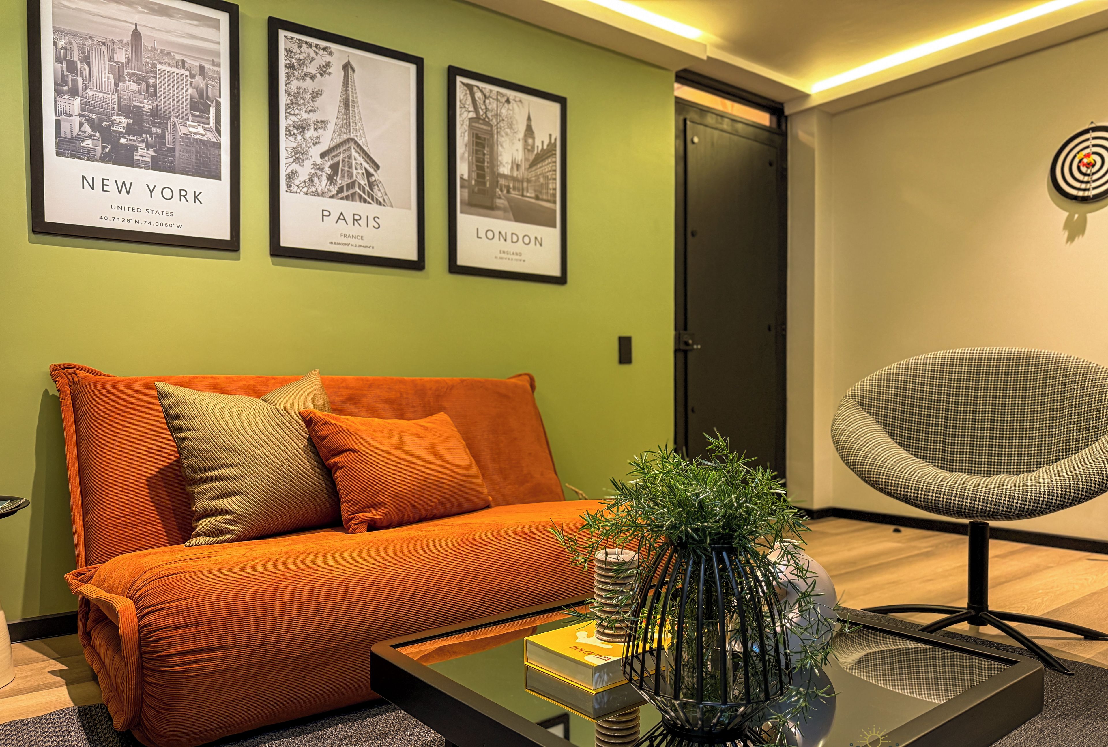 A modern living room with a sofa, a coffee table, and a chair. There are three framed pictures on the wall, one of New York, one of Paris, and one of London.