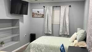 Een slaapkamer met een bed, bureau, stoel, plafondventilator, televisie, radiator en een raam met gordijnen.
