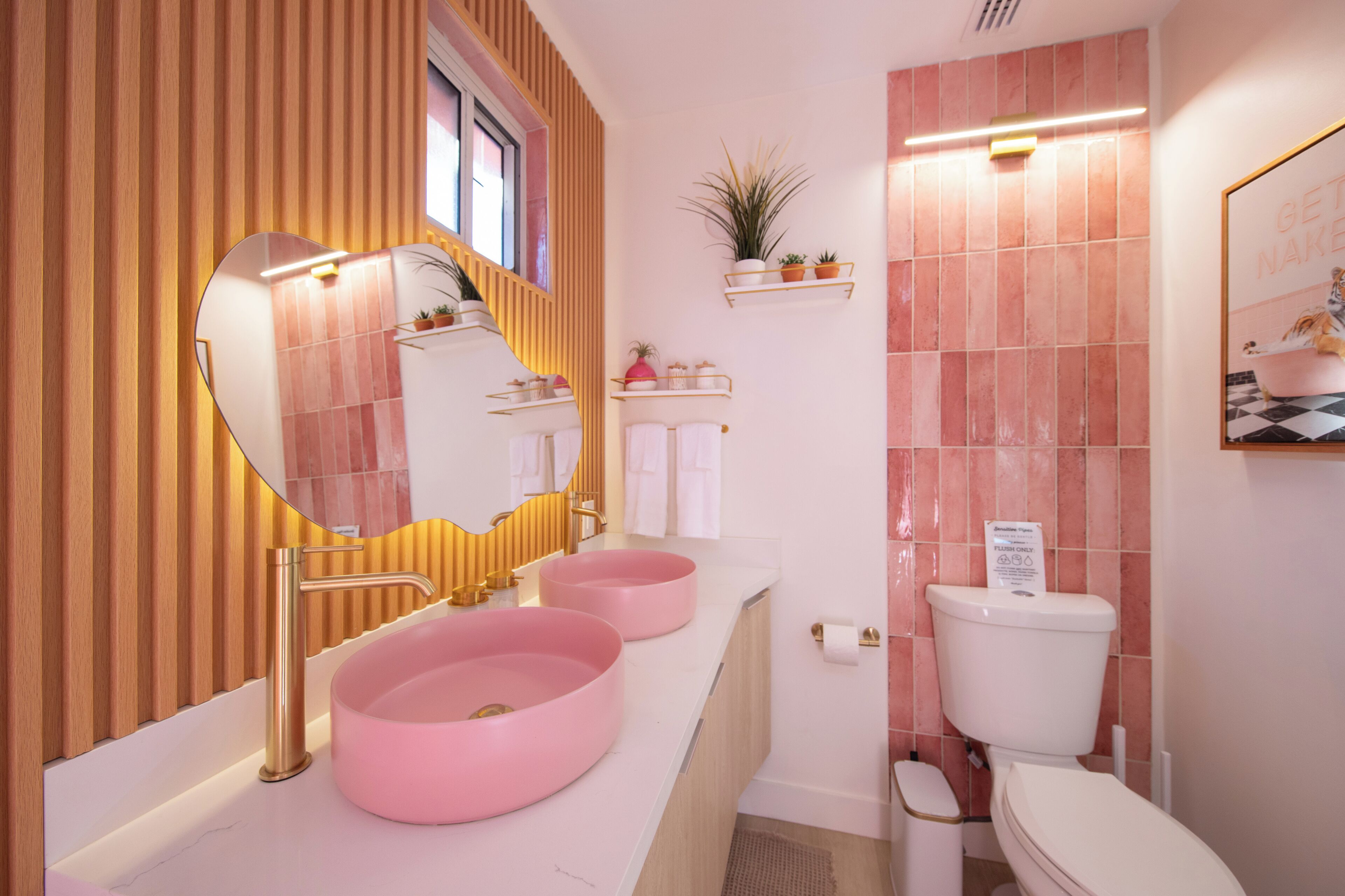 Une salle de bain avec deux lavabos roses, un robinet doré, un miroir, des toilettes et une étagère avec des plantes.