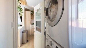 A laundry room with stacked washing machines, a small trash bin, and a door leading to a kitchen area with wooden cabinets and a potted plant.