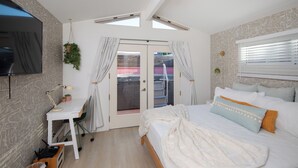 A bedroom with a bed, a desk, a chair, a TV, and a window with blinds.