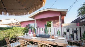 An outdoor dining area with a wooden table and wicker chairs, surrounded by a white picket fence and potted plants.