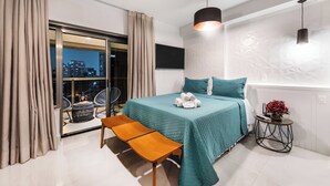 A modern bedroom with a bed, a bench, a small table with a vase, and a view of the city at night.