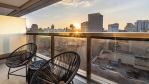 A balcony with two black wicker chairs, a glass railing, and a cityscape view during sunset.