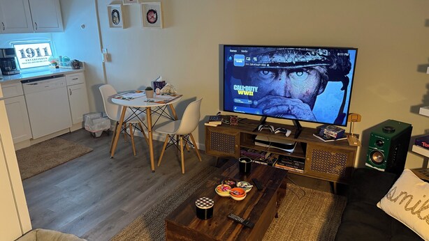A living room with a television displaying a video game, a coffee table with remote controls, a dining area with chairs, and a kitchen area with a coffee machine.