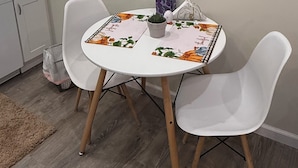 A dining area with a round table, two white chairs, and a floral tablecloth. There are framed artworks on the wall above the table.