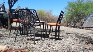 Mobiliário de exterior com cadeiras e mesa de metal, num espaço com gravilha, árvores e um edifício ao fundo.